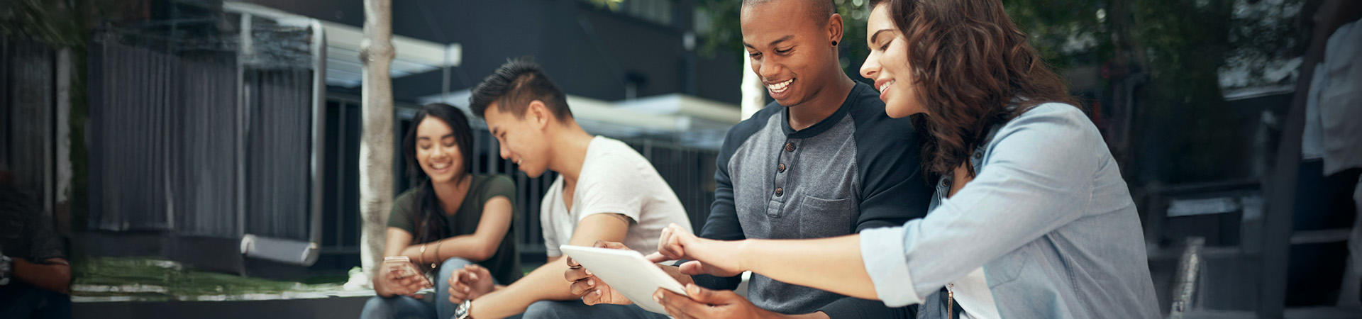 A diverse group of young adults seated on steps, interacting with a tablet, fostering communication and teamwork.